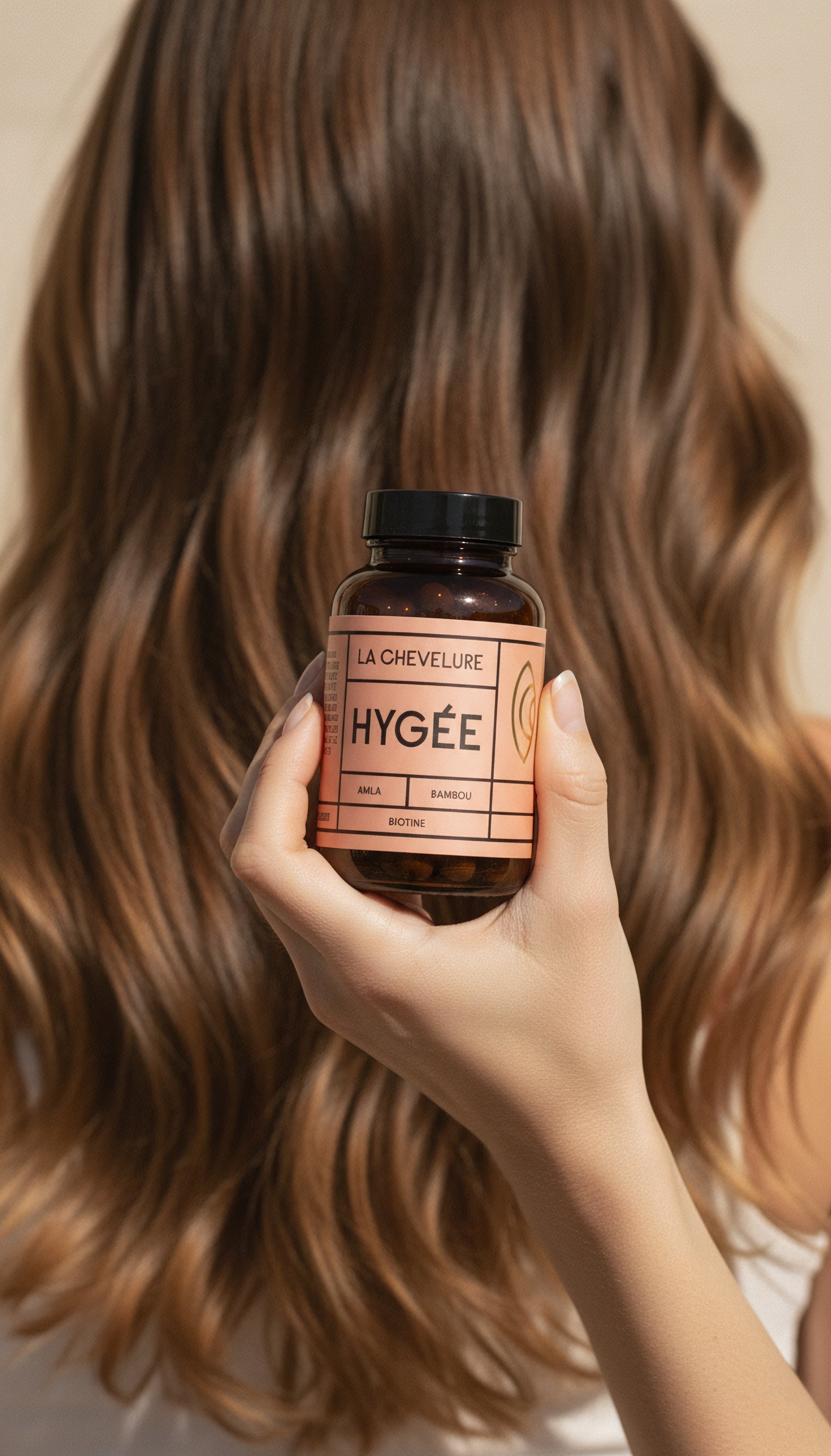 Close‑up of a person holding an amber HYGÉE La Chevelure hair supplement bottle in front of long, wavy, sunlit hair on a warm neutral background.
