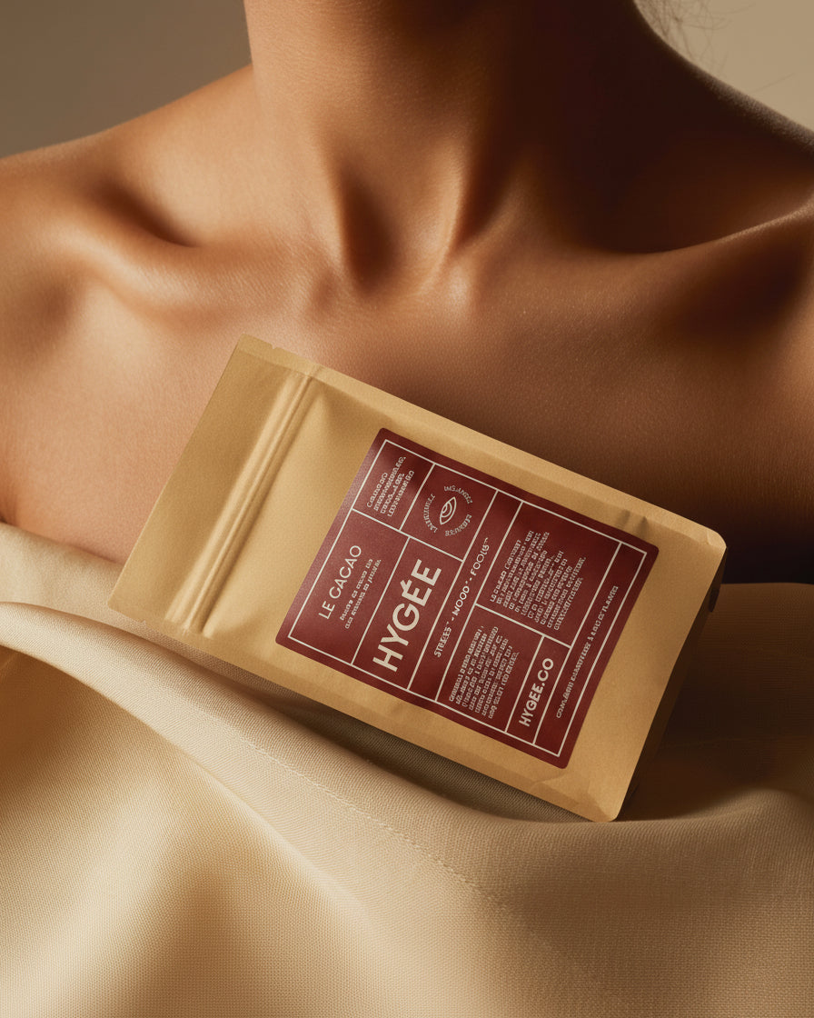 A kraft HYGÉE Le Cacao pouch with a deep burgundy label leans across a collarbone and silky champagne fabric, lit with warm directional light.