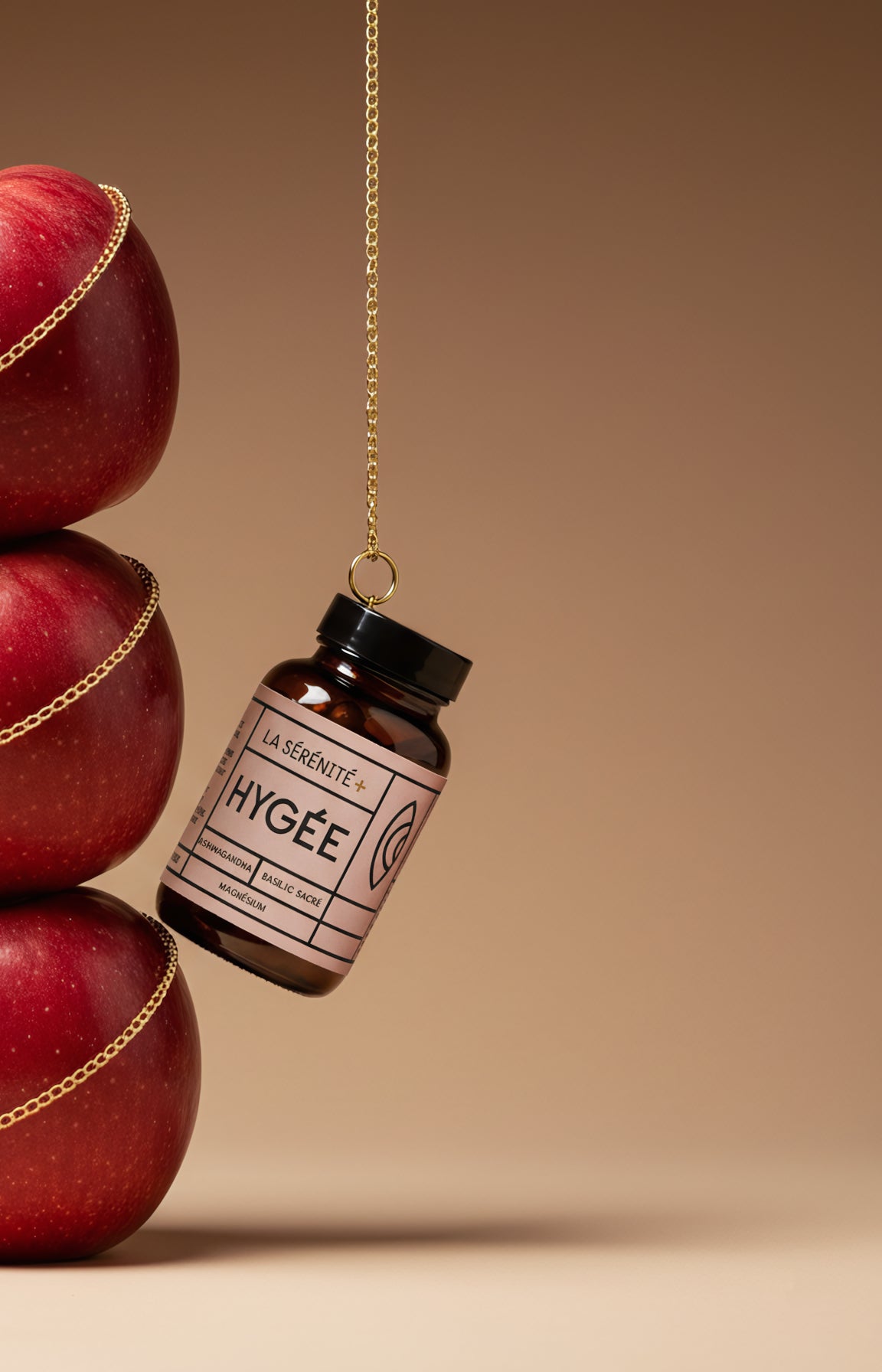 Amber glass bottle of HYGÉE La Sérénité stress‑relief capsules hanging from a delicate gold chain beside a stacked tower of glossy red apples on a warm beige background, creating a luxurious, calming wellness still life