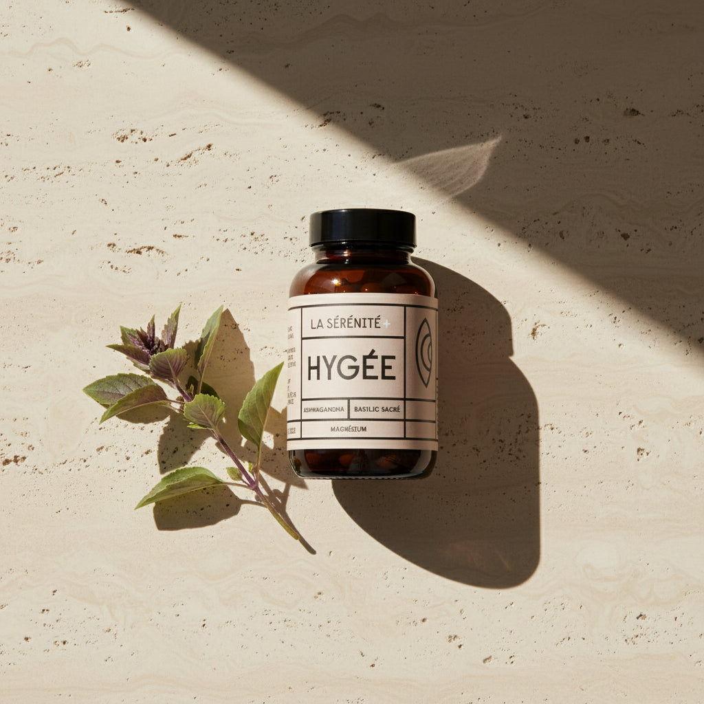 An amber HYGÉE La Sérénité+ bottle with a beige label lies beside a fresh holy basil sprig on a sunlit travertine surface, casting a long diagonal shadow in warm afternoon light.​