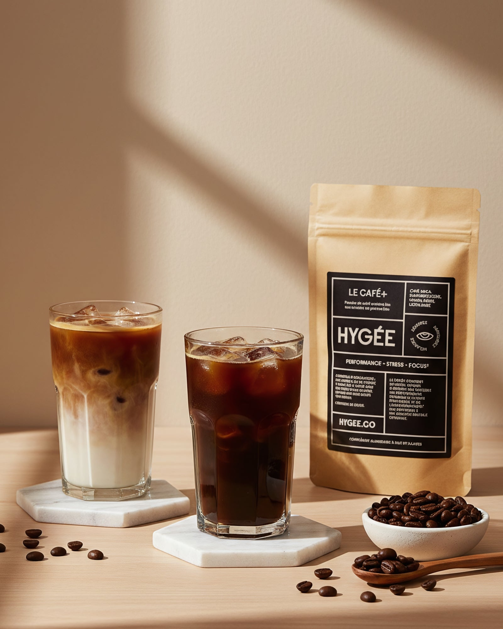 Two tall iced coffees—one layered latte and one black—sit on white hex coasters beside a kraft HYGÉE Le Café+ pouch and a small dish of beans in warm diagonal sunlight. A clean countertop scene with crisp shadows and rich coffee tones.​