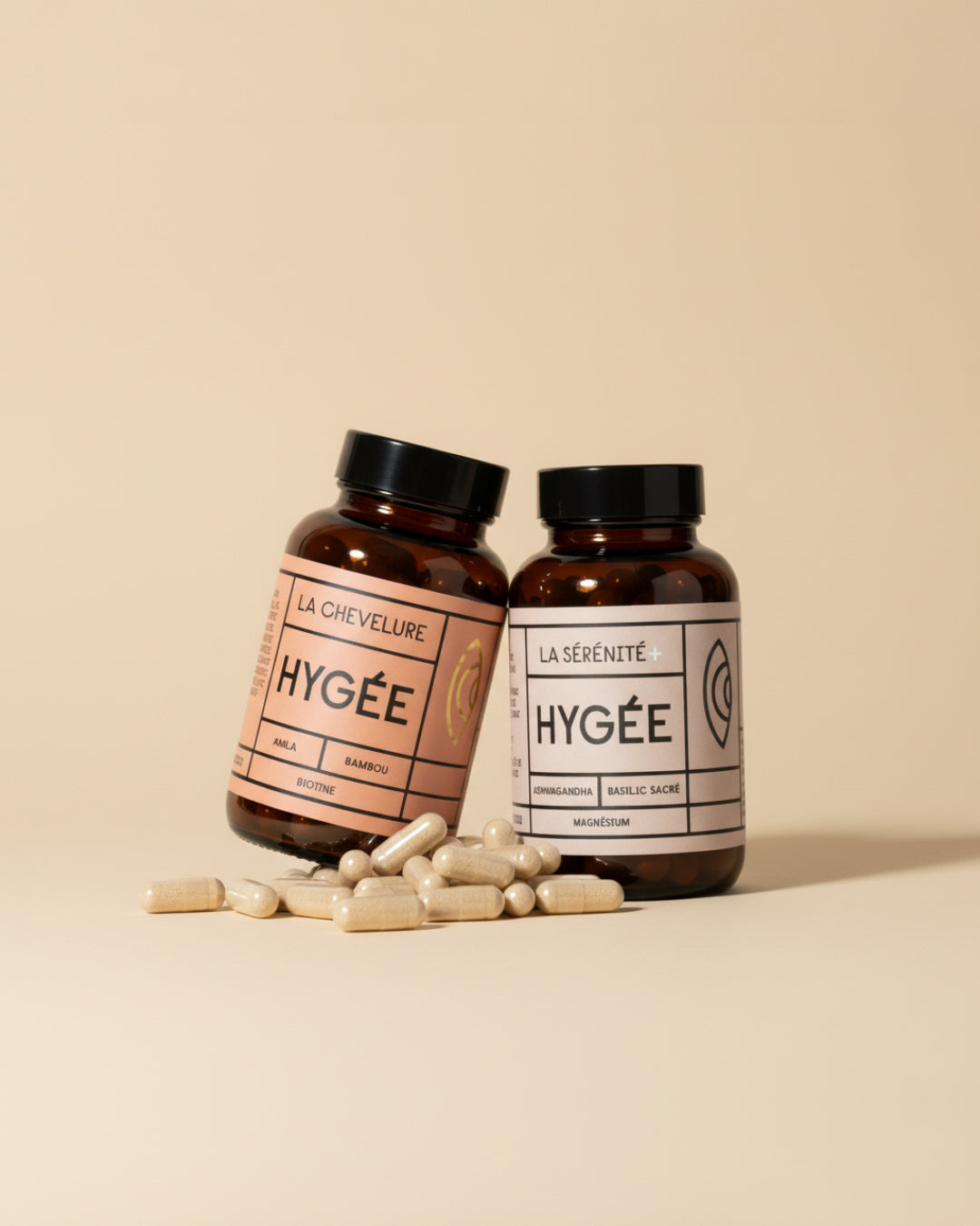 Two amber HYGÉE bottles, La Chevelure and La Sérénité+, leaning together on a soft beige backdrop with a scatter of cream capsules in the foreground, creating a warm minimal wellness scene​.
