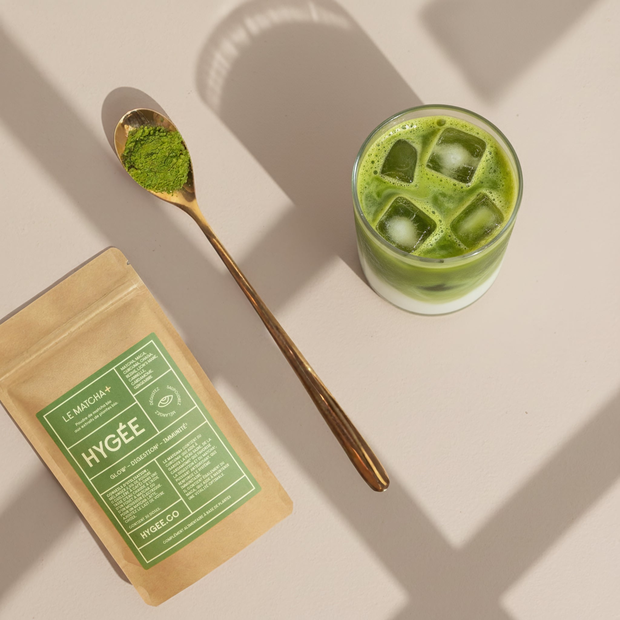 Overhead view of HYGÉE Le Matcha glow and digestion powder pouch beside a golden spoon filled with vibrant green matcha and a layered iced matcha latte on a sunlit neutral surface, highlighting a daily beauty and wellness ritual.​